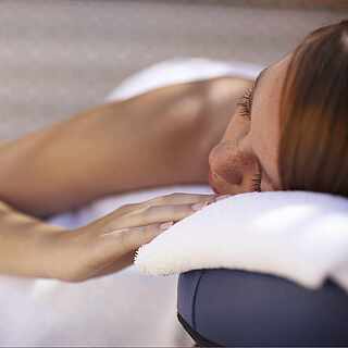 Eine Frau liegt auf einer Massageliege und hat die Augen geschlossen. Sie ist zugedeckt mit weißen Handtüchern.