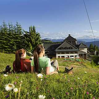 Zwei Wanderinnen liegen auf Bergwiese und schauen auf Berge und Hütte