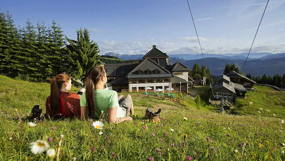 Zwei Wanderinnen liegen auf Bergwiese und schauen auf Berge und Hütte