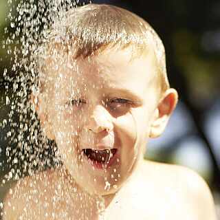 Kleines Kind steht unter einem Wasserstrahler und lacht