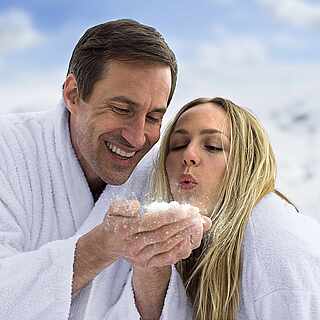 Frau und Mann targen einen Bademantel im Schnee und pusten den Schnee von der Hand des Mannes weg