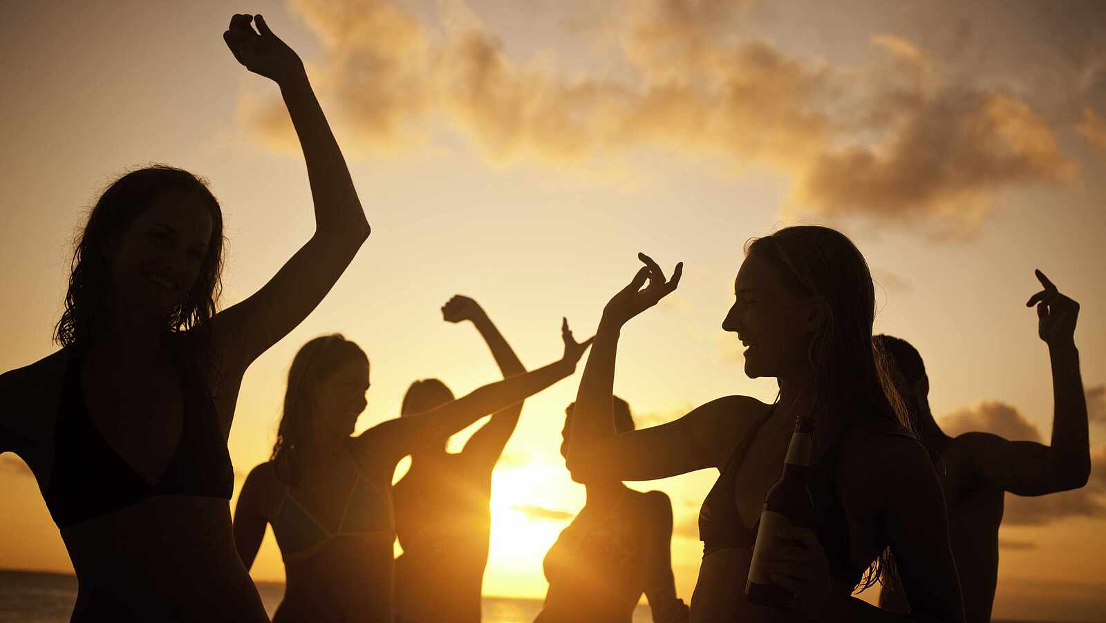 Ein Gruppe von Menschen tanzt beim Sonnenuntergang am Strand, nur die Silhouetten sind zu erkennen.