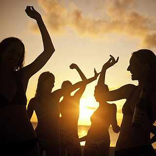 Ein Gruppe von Menschen tanzt beim Sonnenuntergang am Strand, nur die Silhouetten sind zu erkennen.