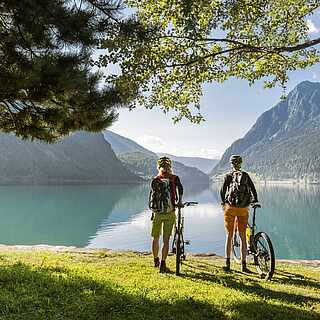 Zwei Personen stehen an einem See. Links und rechts von ihnen stehen Bäume. Der See ist von Gebirgen bzw. Fjorden umgeben.