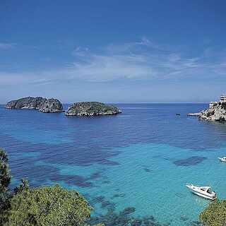 Blick auf klares, türkises Meer mit schroffen Felsen und Booten unter blauem Himmel
