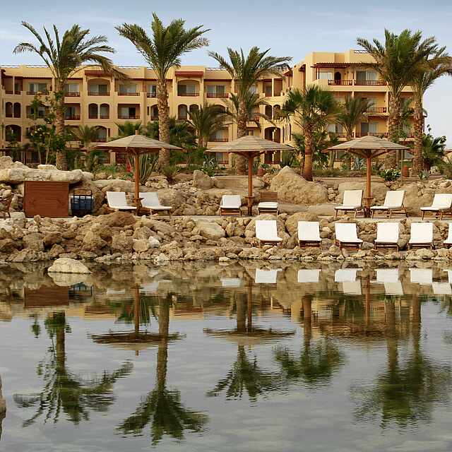 Sonnenliegen an See vor Clubanlage und Palmen