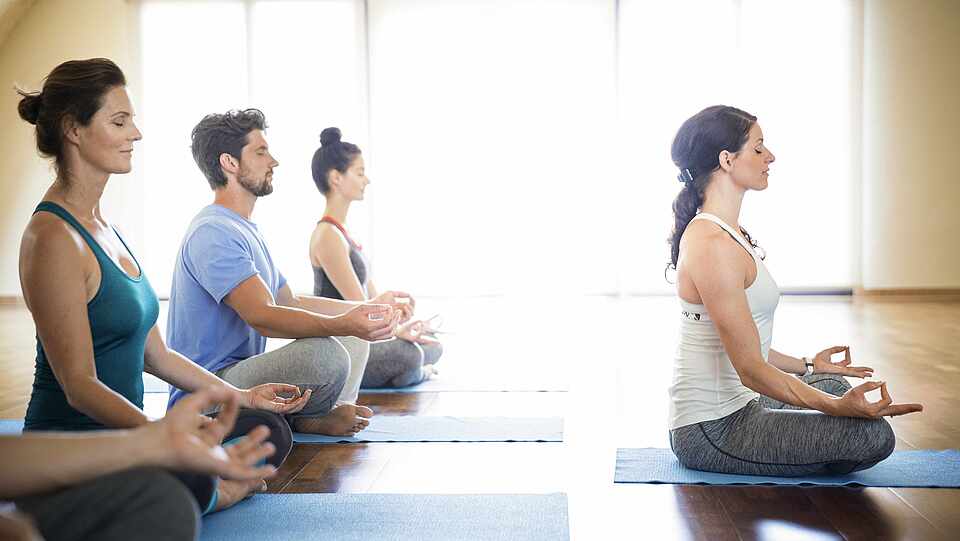 Vier Leute sitzen hinter einer Frau. Alle tragen Sportkleidung und sie sind im Lotussitz.