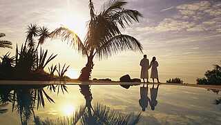 [Translate to English (en_EN):] Paar steht im Sonnenuntergang am Pool mit Palmen