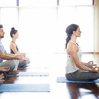 Vier Leute sitzen auf Yogamatten und machen eine Yogaposition. Vor ihnen macht eine Frau die Übung vor.