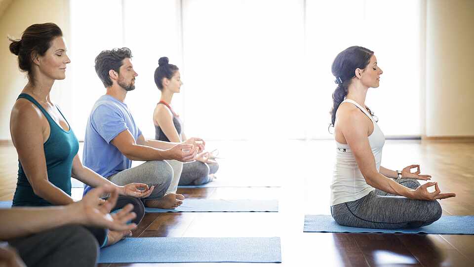 Vier Leute sitzen auf Yogamatten und machen eine Yogaposition. Vor ihnen macht eine Frau die Übung vor.