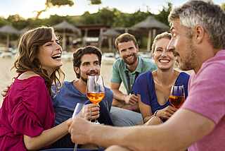 Fünf glückliche Personen die am Strand sitzen und Wein trinken 
