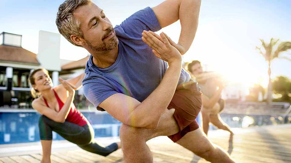 Ein Mann steht vor zwei anderen Personen an einem Pool auf Holz. Alle machen die gleiche Position. Hinter ihnen sieht man links ein Gebäude und rechts Palmen und Liegen.