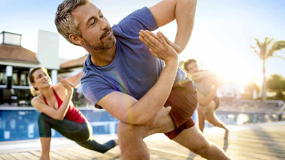 Ein Mann steht vor zwei anderen Personen an einem Pool auf Holz. Alle machen die gleiche Position. Hinter ihnen sieht man links ein Gebäude und rechts Palmen und Liegen.
